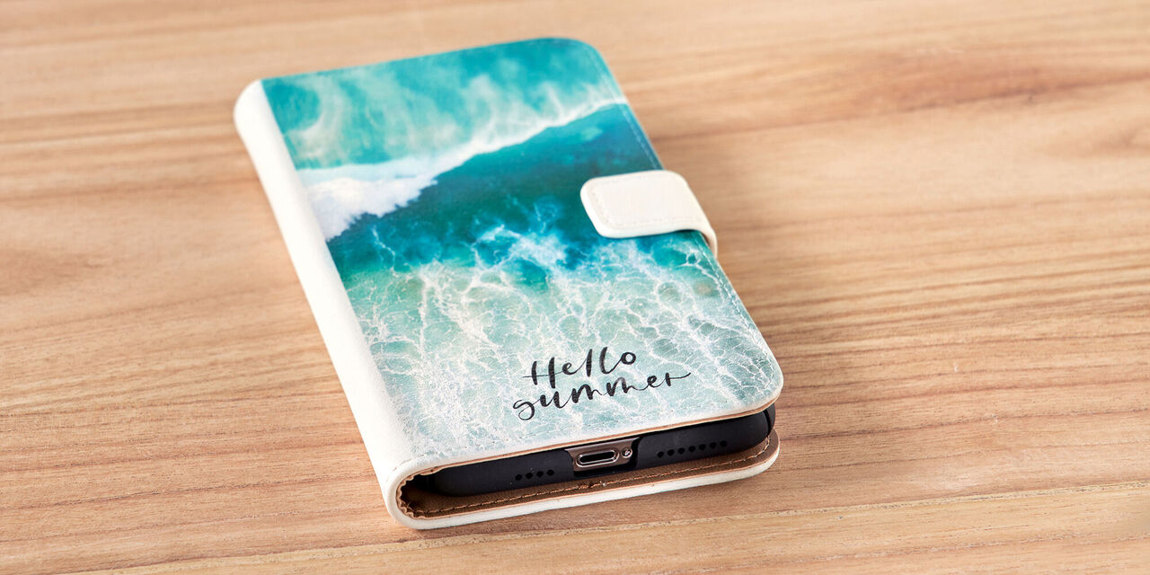 Auf einem Tisch liegt ein Smartphone. Es ist die Handyhülle zu sehen, auf der eine Nahaufnahme von Wasser und die Worte „Hello Summer“ zu sehen sind. Bei der Handyhülle handelt es sich um eine Sideflip Tasche aus weißem Leder