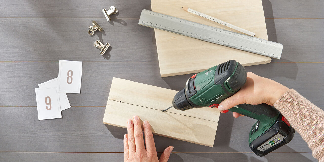 Hände bohren auf einer mit Bleistift gezogenen Mittellinie drei Löcher in ein Holzbrett.