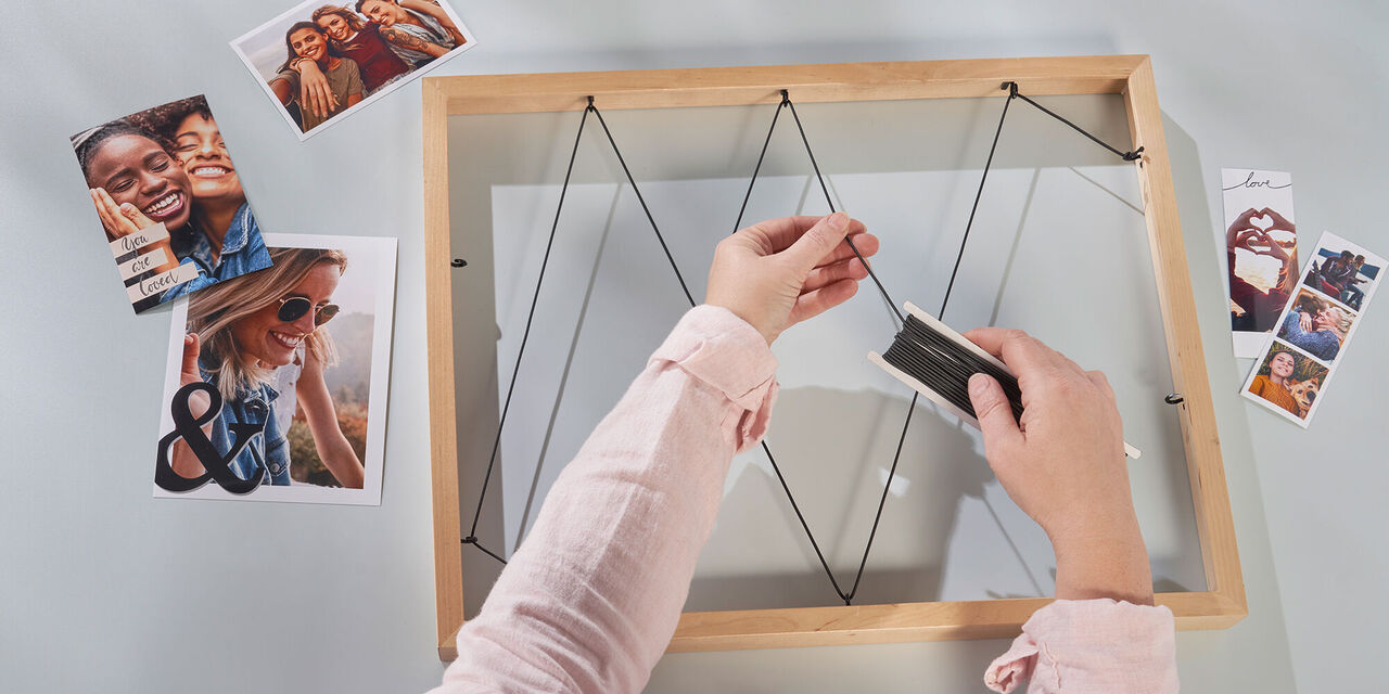 Aufnahme von zwei Händen, die eine Gummischnur um Schraubhaken führen. Diese sind in einen Holzbilderrahmen geschraubt. Neben dem Bilderrahmen liegen Sofortfotos.