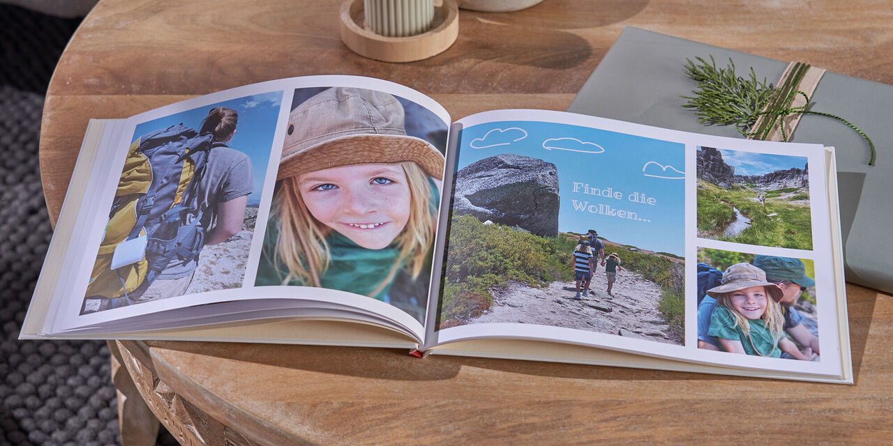 Auf einem Tisch aus Holz liegen ein aufgeschlagenes CEWE FOTOBUCH, zwei Kerzen und ein Geschenk. Auf der Doppelseite sind Fotos von einer Familie beim Wandern zu sehen.