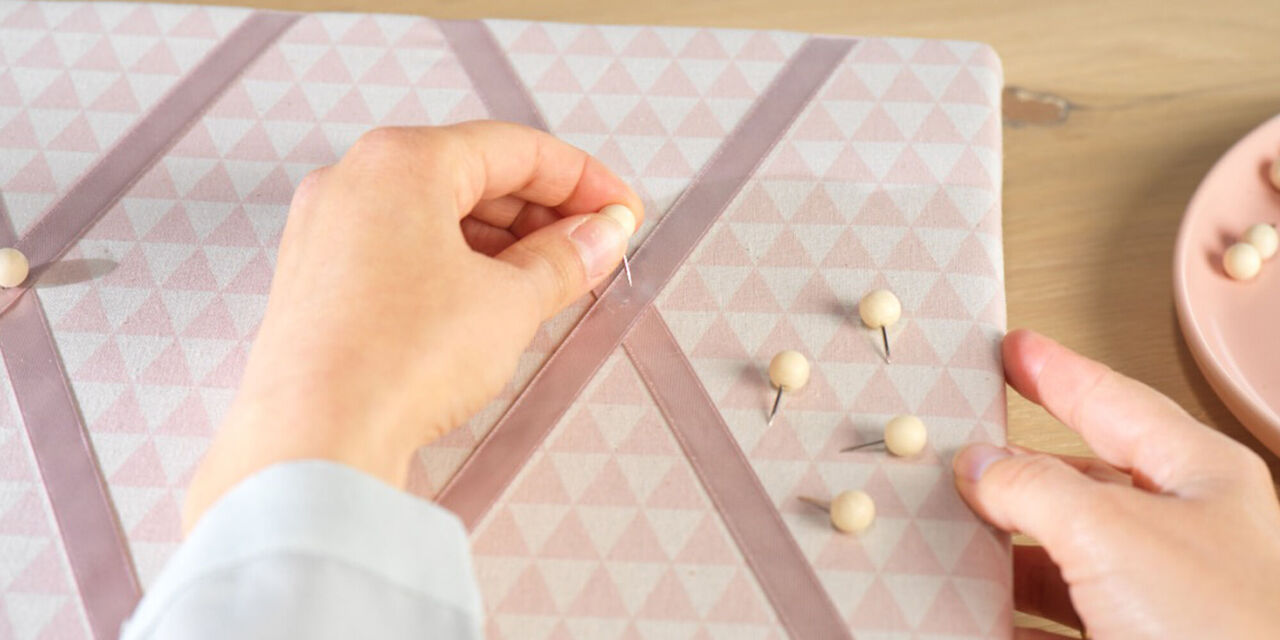 Hand sticht Pinnnadel durch zwei Bänder, die sich auf dem Memo-Board kreuzen.