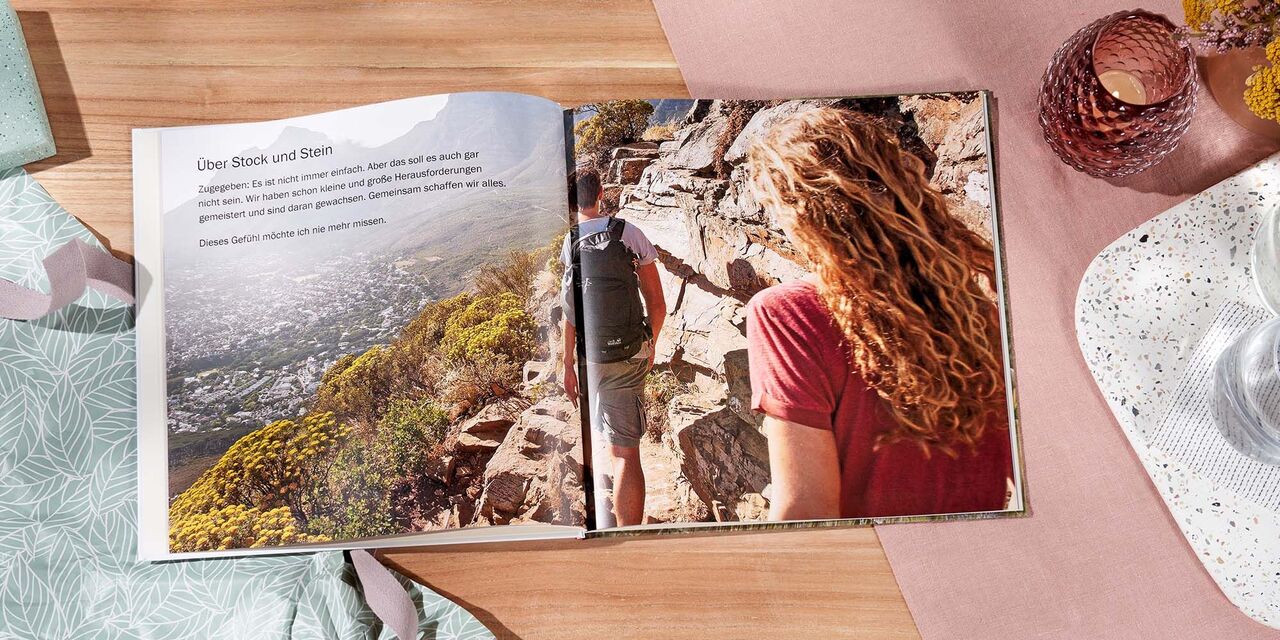 Auf einem Tisch liegt ein aufgeschlagenes CEWE FOTOBUCH. Über beide Seiten erstreckt sich die Aufnahme von einem Paar am Meer. Die Überschrift lautet „Auf die Zukunft“.