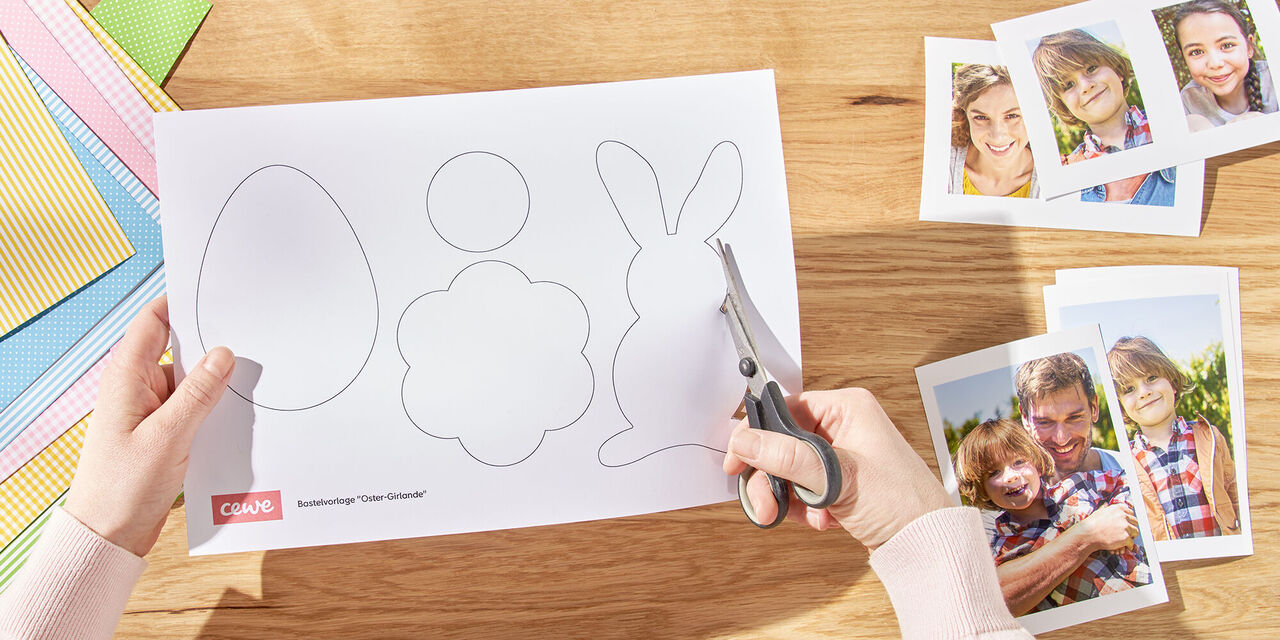 Zwei Hände schneiden einen Osterhasen aus der Vorlage aus. Rechts davon liegen ausgedruckte Sofortfotos der Familie, links liegen verschiedene Bögen Tonpapier.