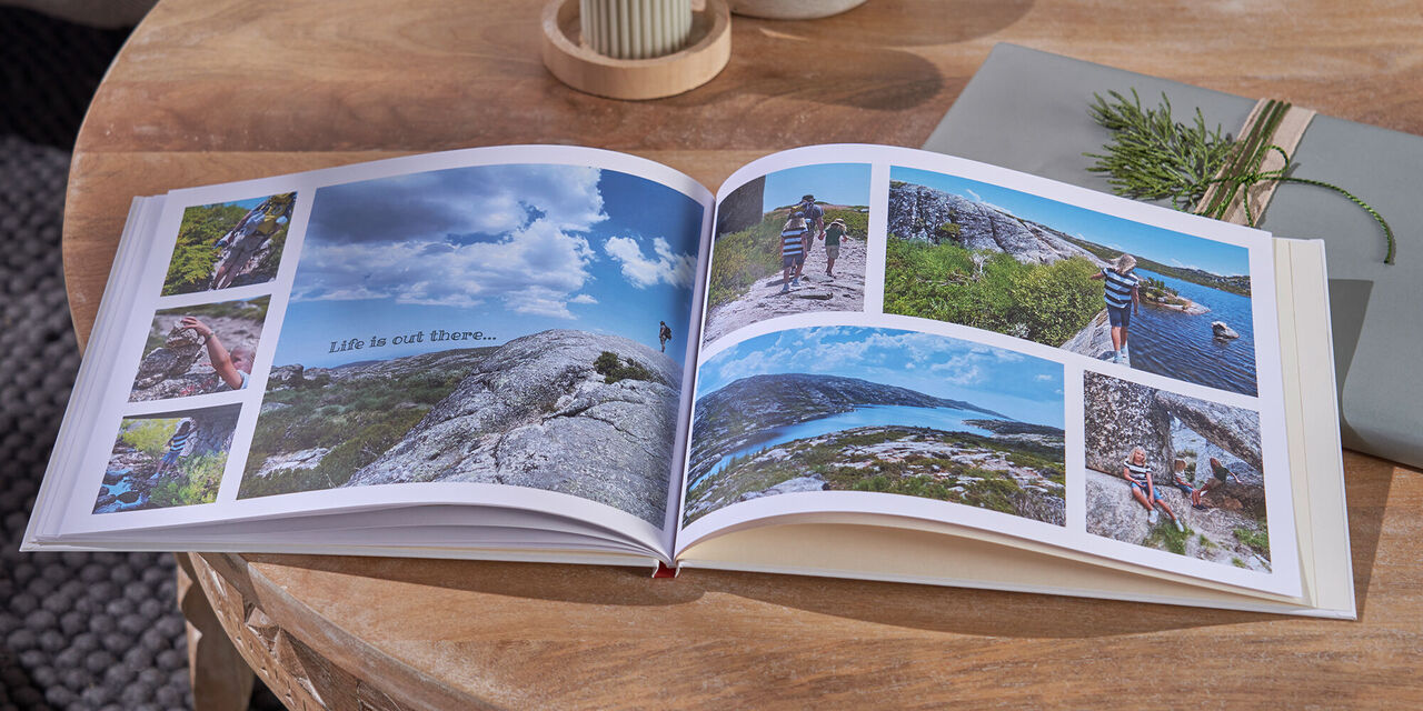 Auf einem Tisch aus Holz liegen ein aufgeschlagenes CEWE FOTOBUCH, zwei Kerzen und ein Geschenk. Auf der Doppelseite sind Fotos von einer Familie beim Wandern in den Bergen zu sehen.